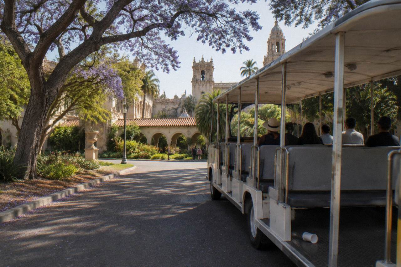 Open-sided park tram on a tree-lined boulevard between gardens and Spanish-style buildings