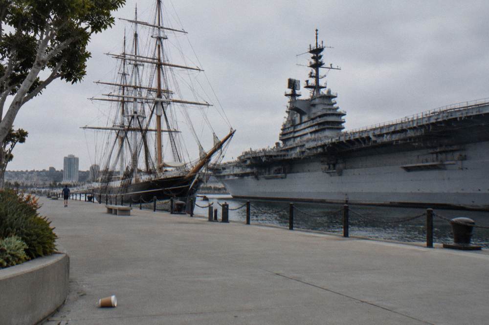A historic sailing ship moored beside a long gray aircraft carrier along a waterfront walkway