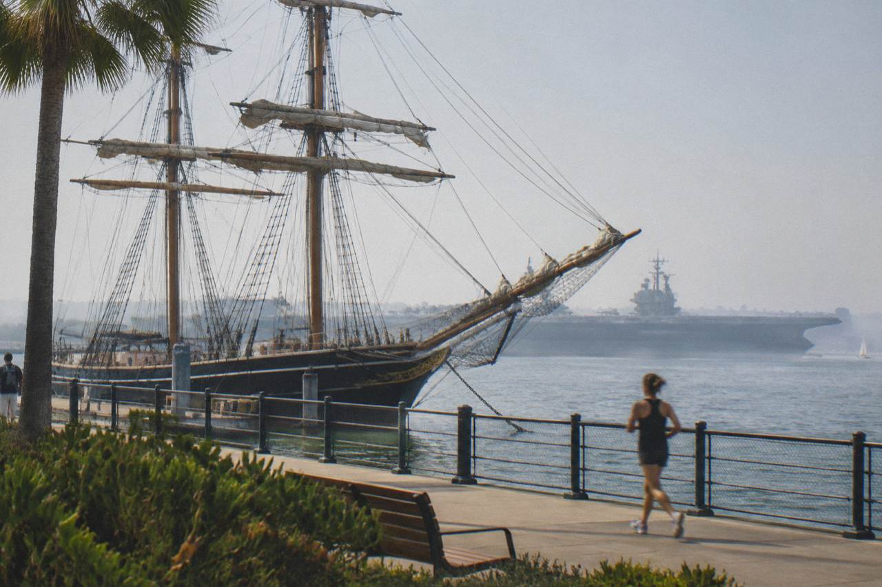A tall sailing ship and a long gray aircraft carrier moored along a waterfront promenade