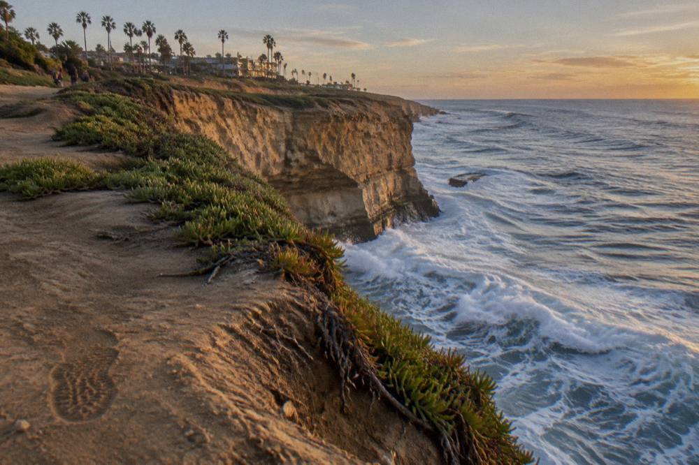 Sandstone cliffs dropping to the ocean with waves breaking along a coastal walking path.