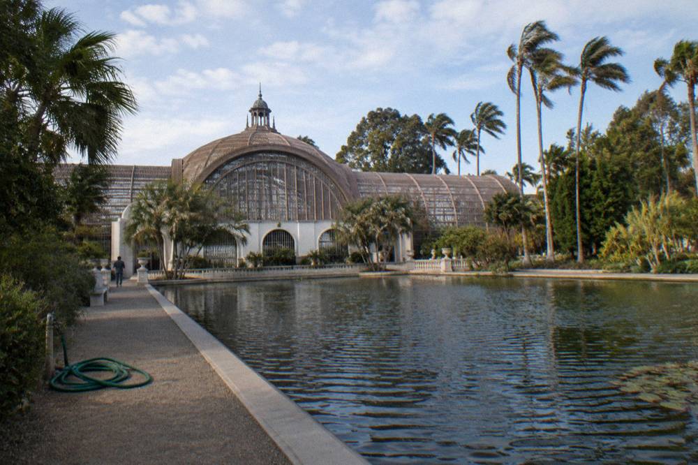 A lath and glass garden building with tall palms and a long reflecting pool in front of it.