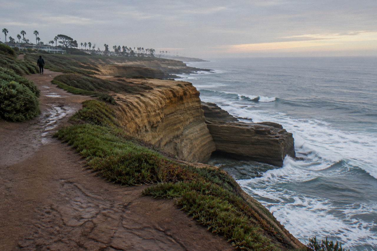 Ocean waves breaking against golden sandstone cliffs along a quiet coastal walking path.