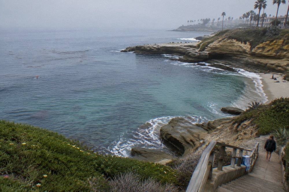 A small curved cove with clear blue water and rocky cliffs along a coastal walkway