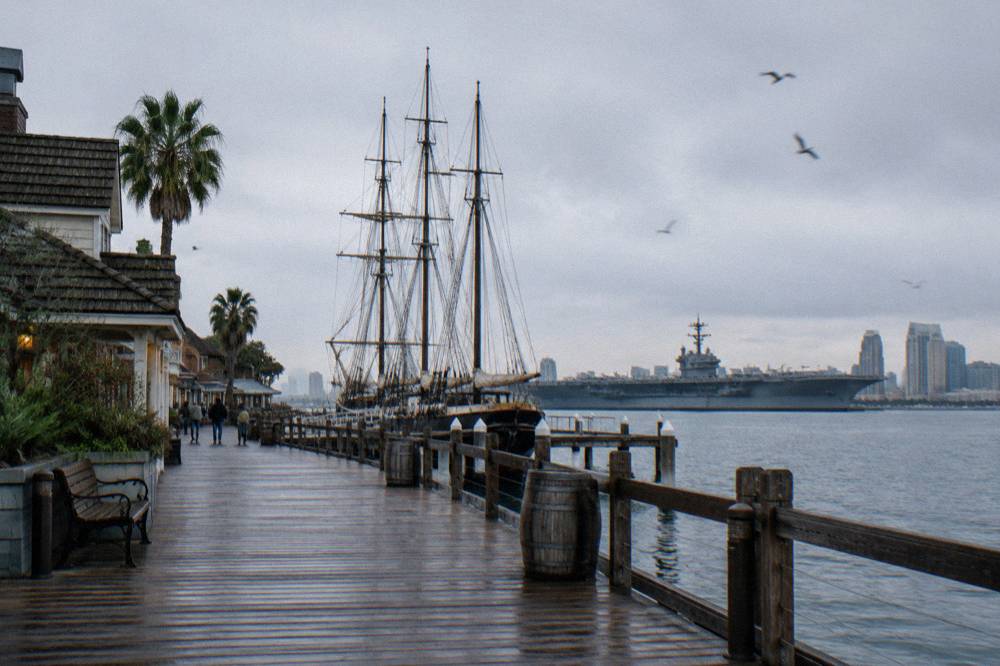 Masts of historic tall ships and a gray aircraft carrier along the San Diego bayfront