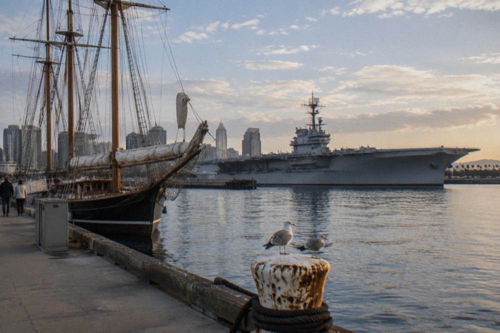 A historic tall ship moored at a harbor pier with a long aircraft carrier visible across the bay