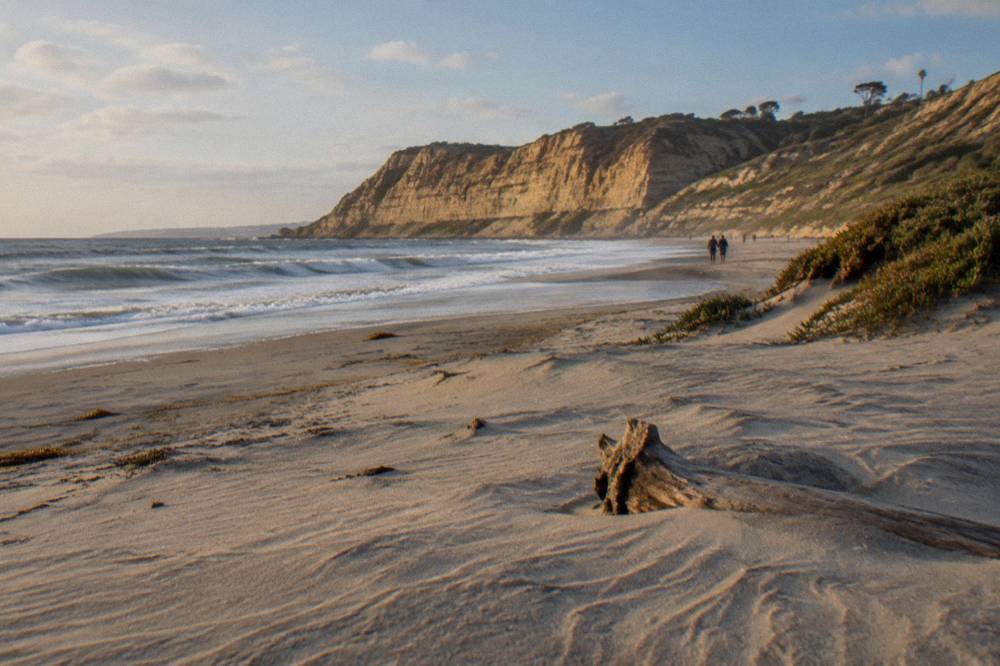 A wide sandy beach with gentle waves and rocky headlands in the distance under a clear sky