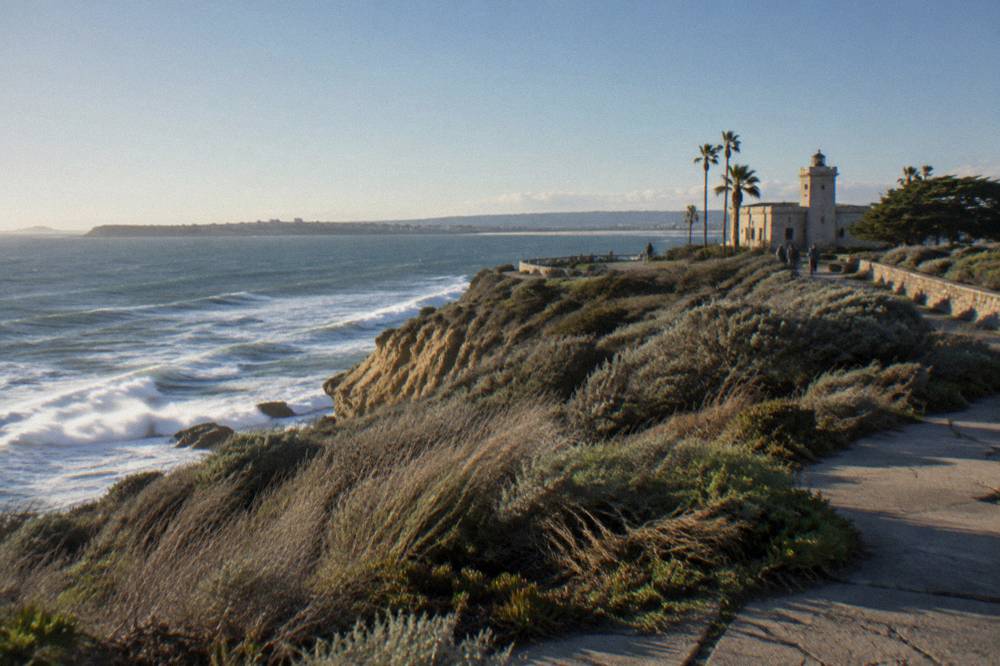 Coastal bluff above the Pacific with waves below and a parking area in the distance
