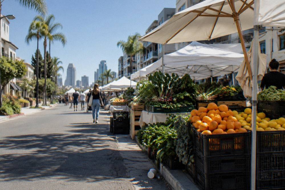 A farmers market street with produce stalls and striped umbrellas along a sidewalk